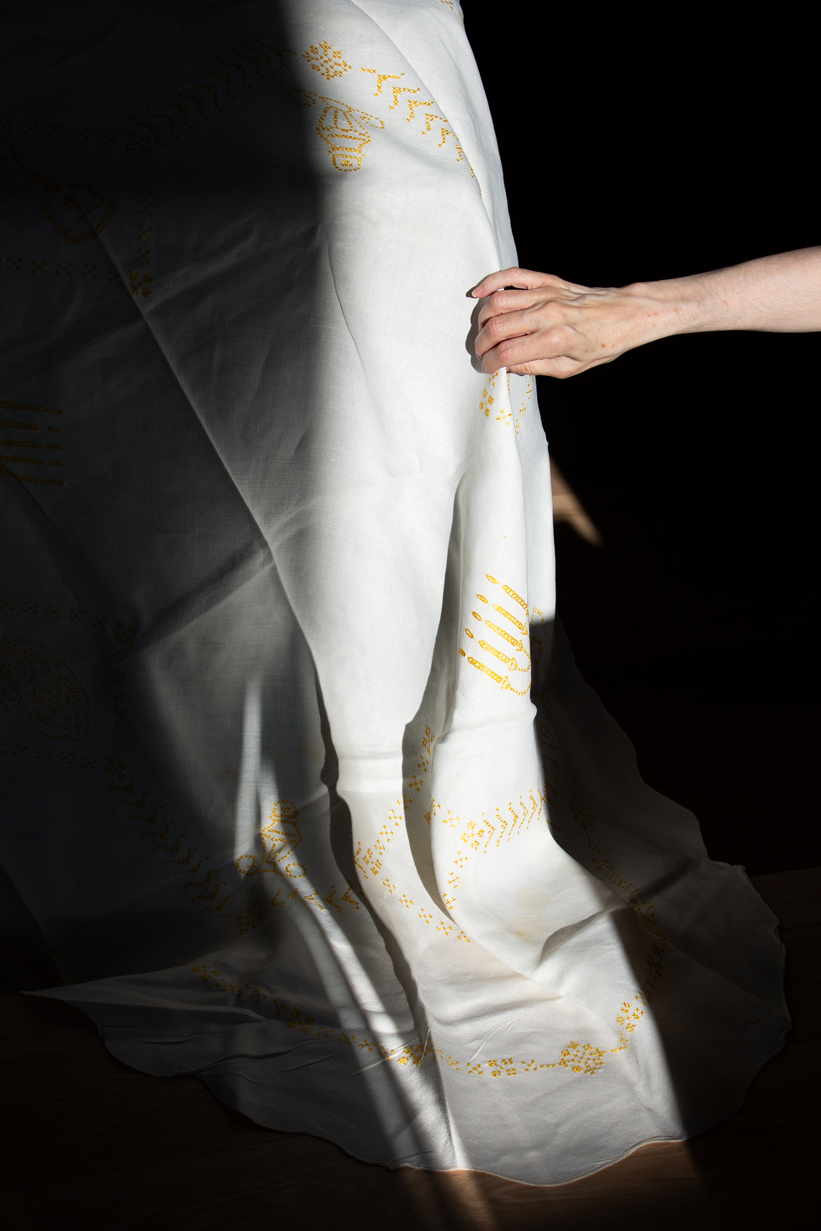 An extended arm reaches out from the right, grasping draped off-white fabric with details embroidered in yellow thread. A bright light is cast over the hand and cloth, contrasted by a deep, dark shadowy ground.