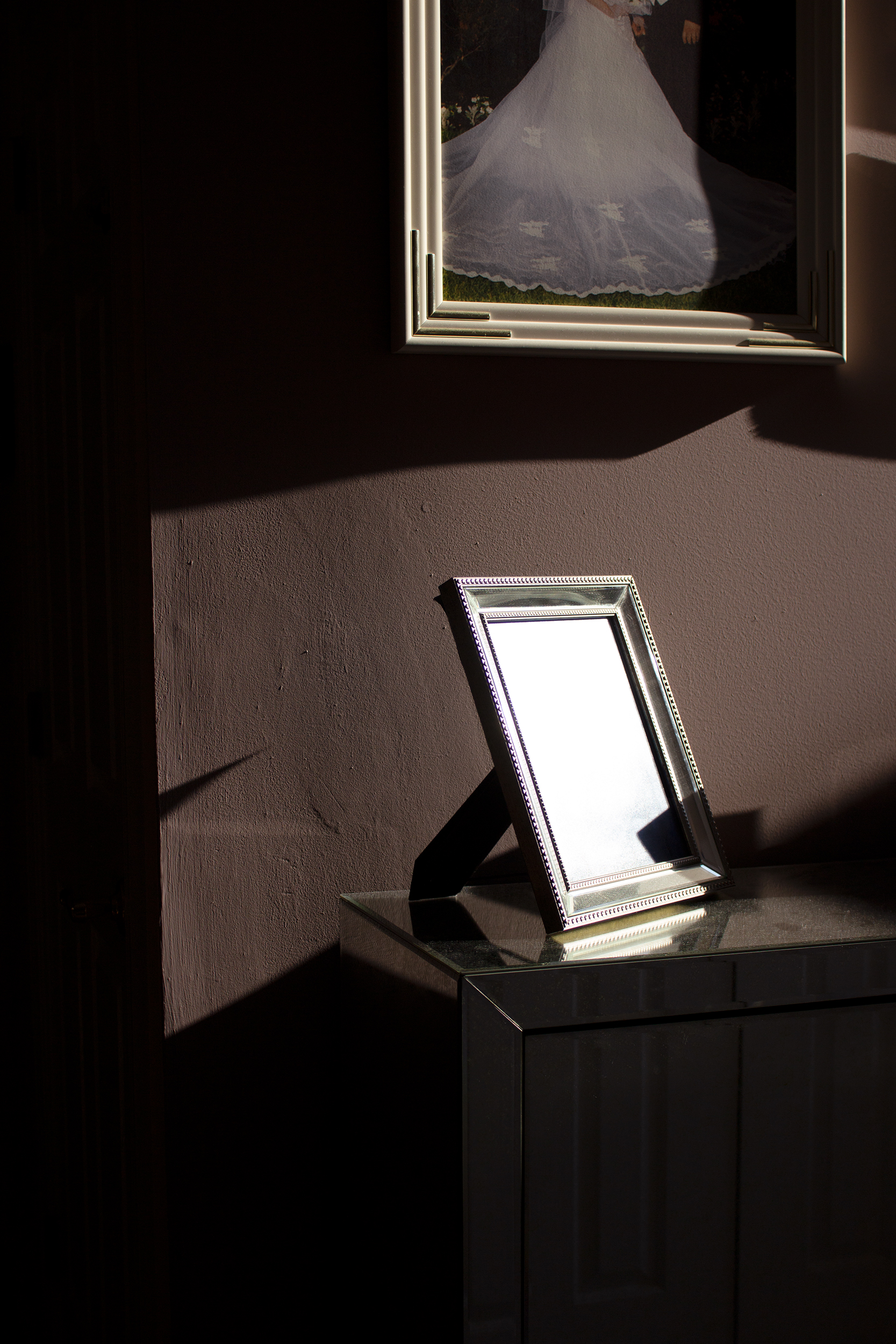 A mirrored picture frame that bright light shines directly on so the picture is filled with light and thus not visible. The frame stands on a mirrored corner of furniture, in front of a mauve wall cast with dramatic shadow contrasted by bright light cutting through the center. On the wall above hangs a framed wedding photo, with only the bottom half of two figures visible.